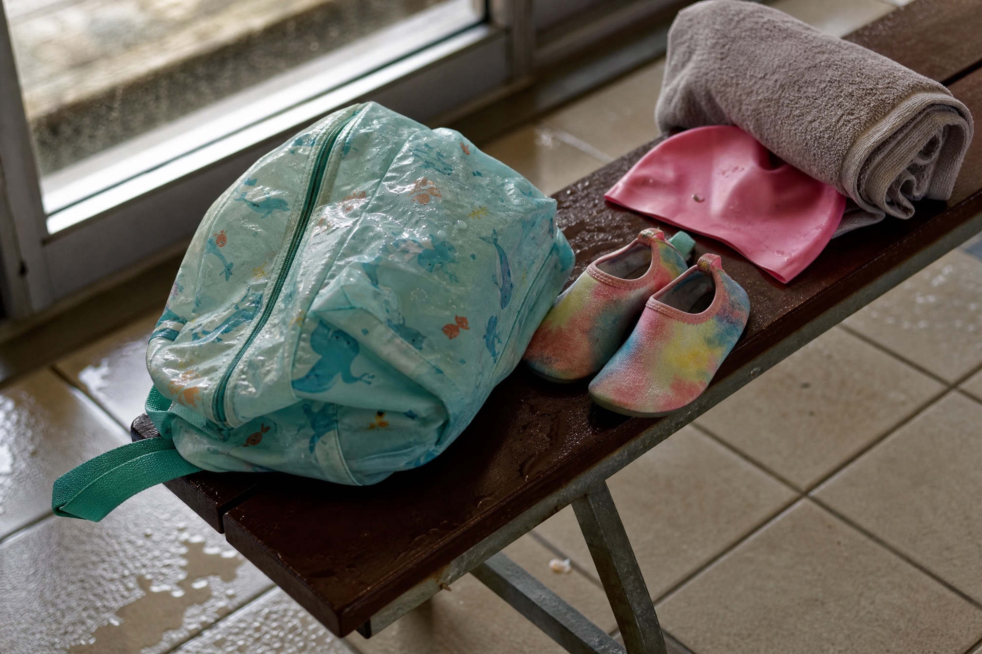 A childs swim bag, water shoes, swim cap and towel laid out on a pool-side bench before a Dutch swimming lesson