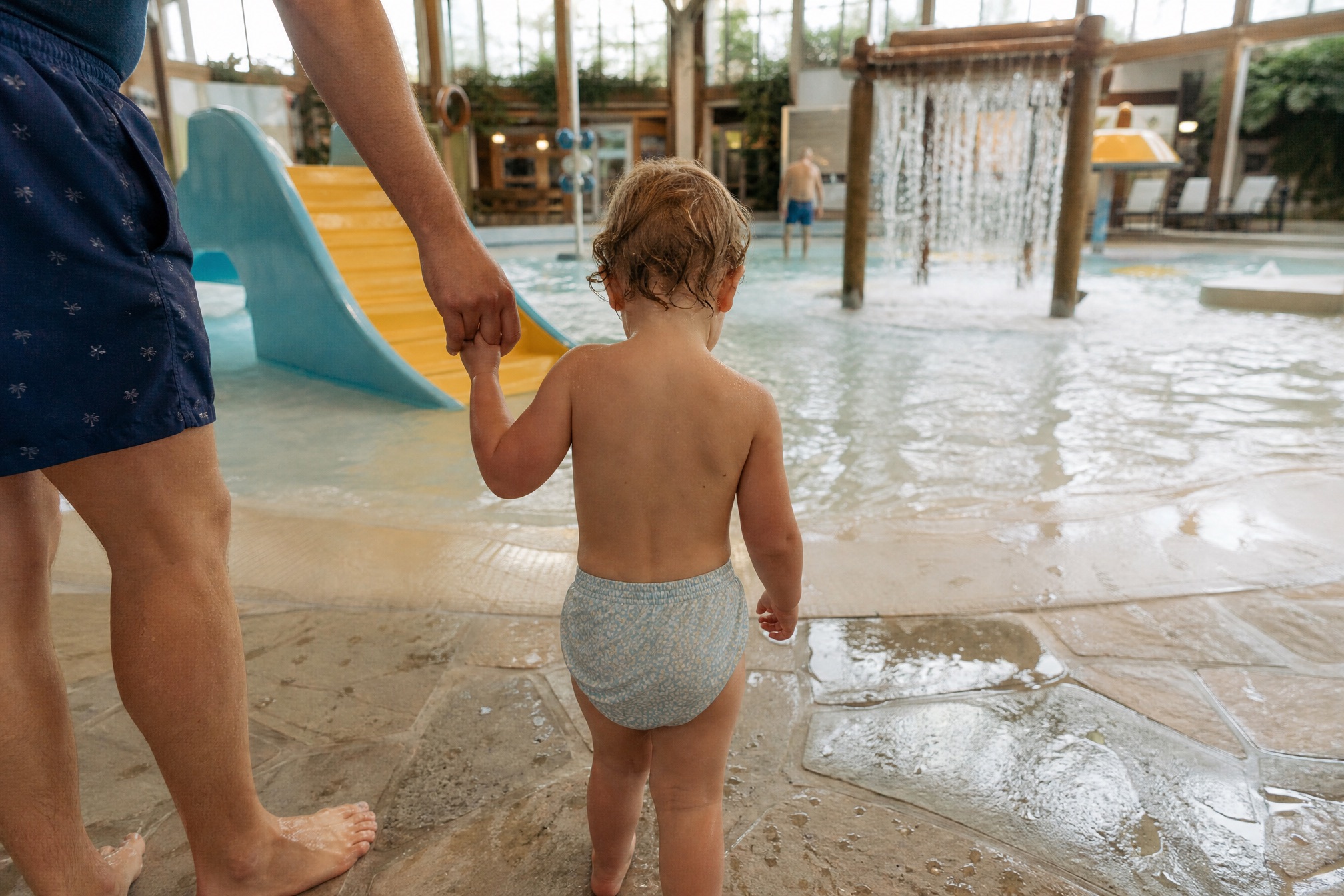 Toddler holding a parent hand next to a small toddler water slide indoors