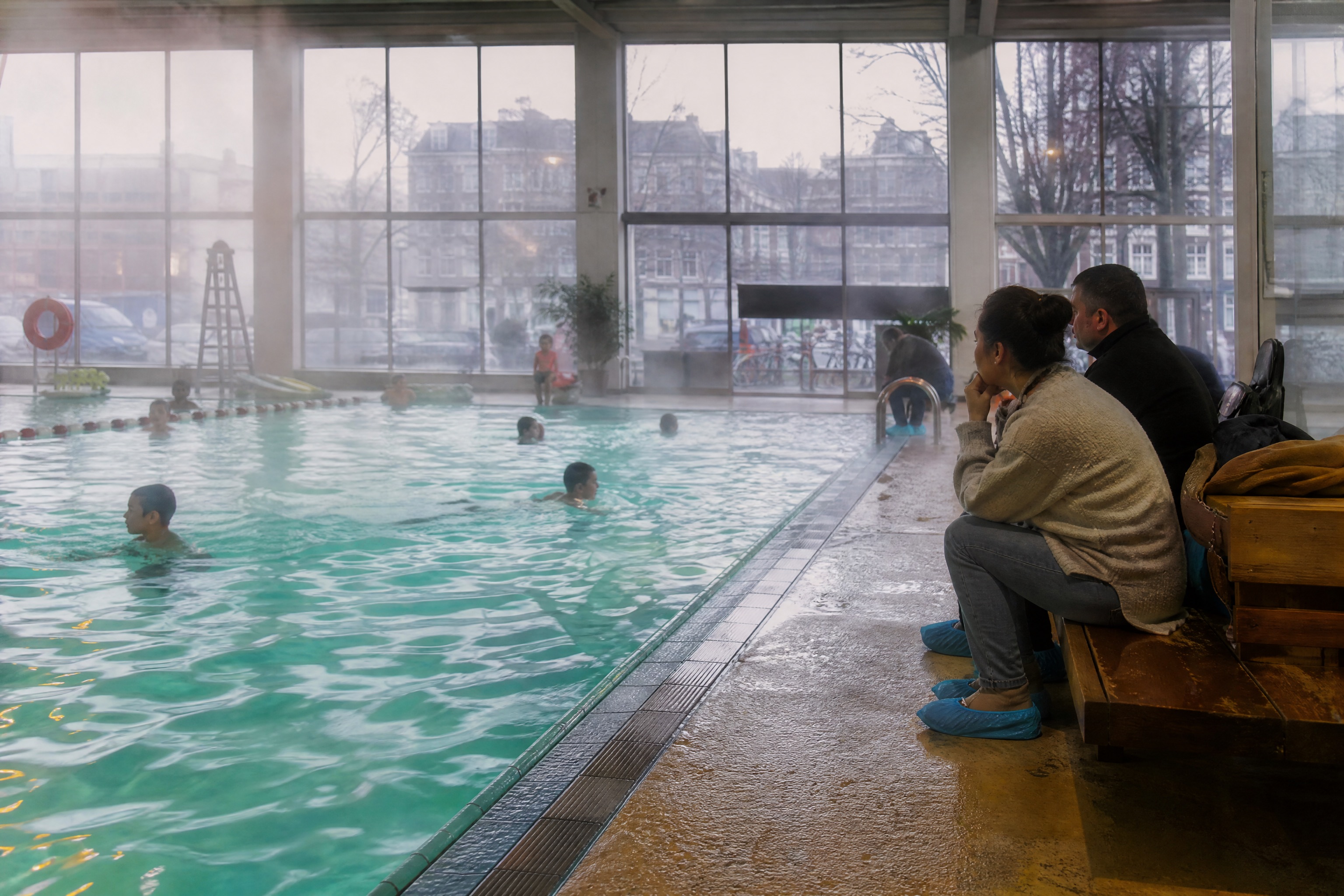 Parents sitting poolside at an indoor Amsterdam swimming pool on a grey rainy day