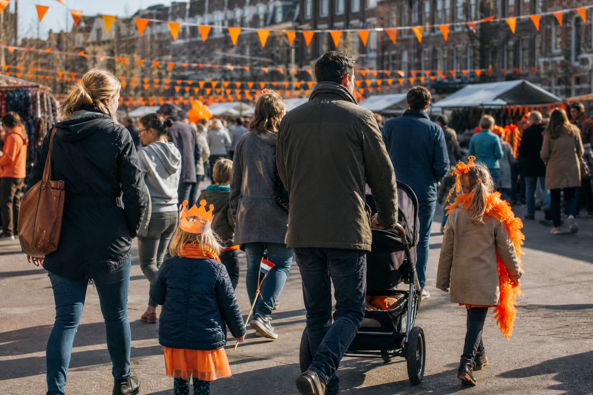Family pushing a stroller through an orange-decorated street market on Kings Day 2027